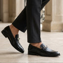 A person wearing black leather penny loafers with a small silver horsebit detail, paired with dark charcoal gray trousers, walking on a light beige stone terrace.