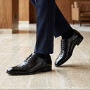 Person wearing black dress shoes and navy pants in a room with wooden flooring and a blurred background.