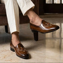 Man sitting on a brown armchair wearing a navy jacket, white pants, and brown shoes in a room with a bookshelf.