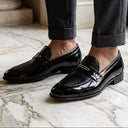 A person wearing black patent leather loafers with gold-tone bit details, paired with dark gray cuffed trousers, standing on a polished marble floor.