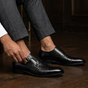 Man adjusting his black leather shoes in a room with wooden floor and furniture.