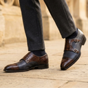 A close-up of a person's legs wearing dark gray trousers and brown double monk-style dress shoes with a mix of smooth leather and crocodile-embossed textures, standing on a stone pavement.