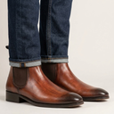 A pair of men's brown leather Chelsea boots worn with dark blue cuffed jeans, shown from a side profile against a light off-white background.