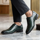 A person wearing dark charcoal gray trousers and glossy dark green crocodile-embossed oxford shoes, standing on a polished marble floor in an indoor setting.