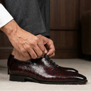 A man in a white dress shirt and gray trousers is crouching down to tie the laces of a pair of dark burgundy crocodile-embossed leather dress shoes in a home office setting.