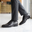 A person wearing dark gray dress pants and black crocodile-embossed leather oxford shoes, standing on a polished marble floor in a modern office lobby.