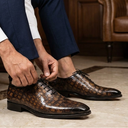 A man wearing a white dress shirt and navy trousers sits on a brown leather sofa, tying the laces of his brown crocodile-embossed leather dress shoes in a warm, wood-paneled room.