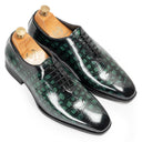 A pair of men's dress shoes with a glossy green and black crocodile-embossed leather finish, displayed on wooden shoe trees against a white background.