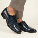 A pair of men's dark blue crocodile-embossed leather oxford shoes worn with gray trousers, shown against a plain off-white studio background.
