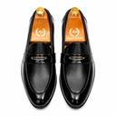 A pair of black leather loafers shown from a top-down view on wooden shoe trees. The shoes feature a glossy patent leather toe cap, a textured chevron-patterned vamp, and a black leather strap with a gold-tone metal logo plate.