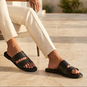 Man sitting outdoors wearing black sandals and light-colored pants.
