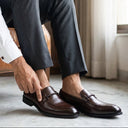 Man adjusting his brown leather shoes in a room with a brown leather chair and light-colored walls.