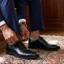 Man in a blue suit adjusting his black shoes in a room with wooden furniture and books.