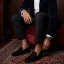 Man in a blue velvet suit sitting on a red stool in a room with wooden walls.
