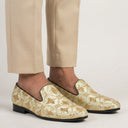 Gold patterned loafers worn with beige pants on a light background