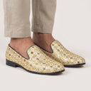 Gold patterned loafers worn with beige pants on a white background