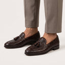 Brown leather loafers with tassels worn with gray pants on a white background