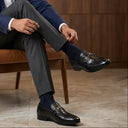 Man in a blue blazer and gray pants sitting on a brown leather chair.