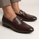 Brown leather loafers with gold buckles worn by a person on a light background