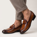 Brown loafers with tassels worn with beige pants on a neutral background