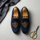 Pair of navy velvet slippers with gold embroidery on a gray surface, accompanied by a pair of gold scissors.