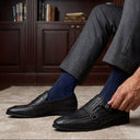 Man in gray suit and blue socks adjusting black loafers in a room with bookshelves.