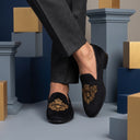 Black loafers with gold embroidery worn by a person, set against a geometric background.