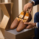 Brown woven loafers being held by a person wearing a suit, with a wooden shoe tree in the background.
