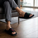 Man sitting on a leather chair wearing black slides with green and red accents. home slippers.