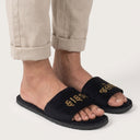 Black slides with gold text worn with beige pants on a white background. velvet emrboidery slogan language slippers for home comfort men.
