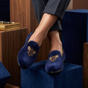 Person wearing blue velvet slippers with gold embroidery on a blue surface.