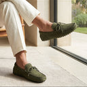 Man sitting on a chair wearing green loafers and white pants, with a window in the background.