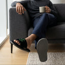 Man sitting on a couch holding a mug, wearing black slides with green accents.