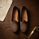 Pair of brown loafers with gold emblem on a wooden surface with a tape measure and paper. wedding juttis