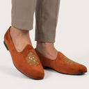 Brown suede shoes with gold embroidery worn with beige pants on a white background. wedding punjabi jutti with zardozi.