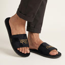Black sandals with gold text worn by a person on a neutral background. kya challa logan home slippers