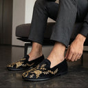 Man wearing black loafers with gold embroidery sitting on a gray sofa. zardozi wedding festive shoes for men.