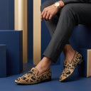 Person wearing patterned loafers and a watch, sitting on a blue block with a blue background.