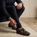 Man sitting on a couch wearing black embroidered shoes in a room with a neutral color palette.