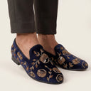 Navy velvet slippers with gold floral embroidery worn with black pants on a neutral background