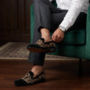 Person sitting on a green chair wearing black loafers with gold embroidery, white shirt, and gray pants.