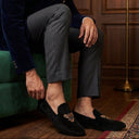 Man sitting on a green chair wearing a navy velvet jacket and gray pants, adjusting his black loafers with a gold emblem.