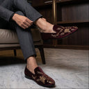 Man sitting in a chair wearing gray suit and burgundy loafers with gold embroidery.