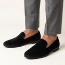Black loafers worn with gray trousers on a white background. velvet slipons