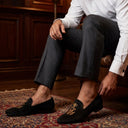 Man sitting on a leather chair wearing black loafers in a room with wooden paneling and a lamp.