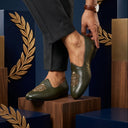 Green shoes with intricate designs worn by a person, set against a decorative background with laurel wreaths.