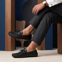 Person wearing black loafers and dark trousers sitting on a wooden stool.