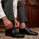 Man in formal attire adjusting his black shoes in a room with bookshelves.