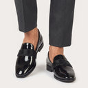 Black patent leather loafers worn with gray trousers on a light gray background
